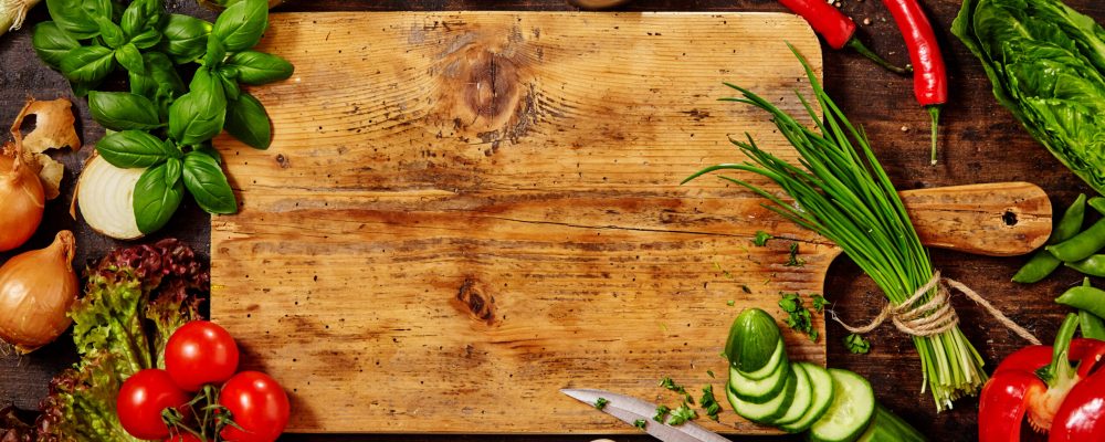High Angle Still Life View of Knife and Wooden Cutting Board Surrounded by Fresh Herbs and Assortment of Raw Vegetables on Rustic Wood Table