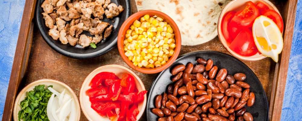 Ingredients for Mexican food on a tray: beans, stew beef, corn, tomatoes, red pepper, onion, herbs, tortillas taco, top view copy space
