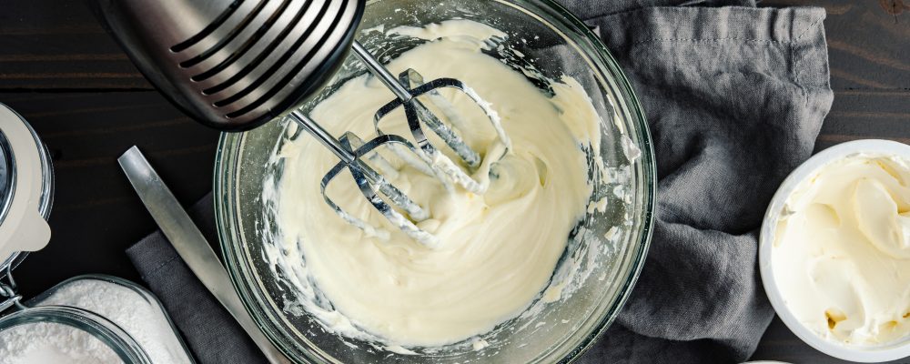 Homemade Cream Cheese Frosting with hand Mixer: Glass mixing bowl of beaten heavy whipping cream, mascarpone cream, and powdered sugar