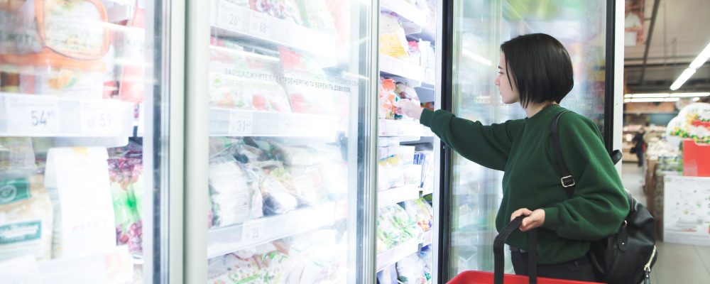 A girl with a basket takes frozen foods from the supermarket's refrigerator. The girl chooses goods in the store. Shopping in the supermarket