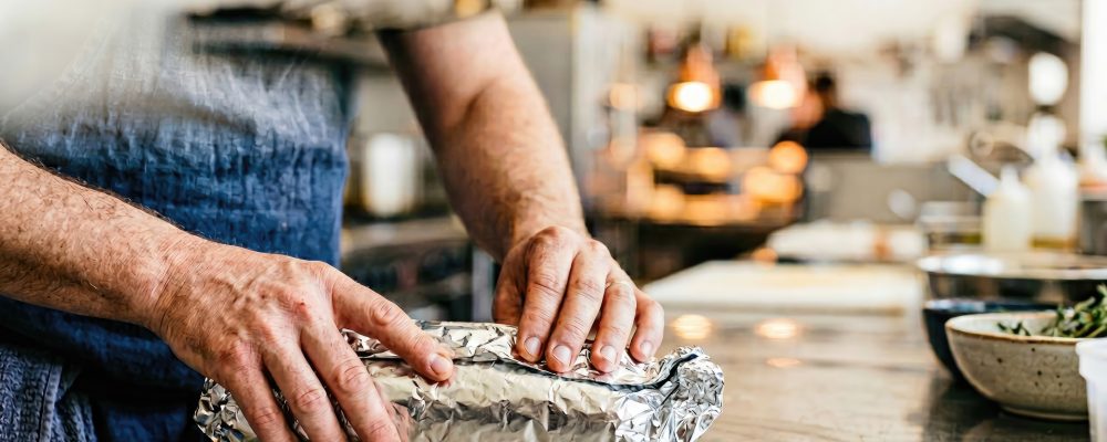 A chef wrapping a burrito or sandwich in aluminum foil on a stainless steel counter in a kitchen