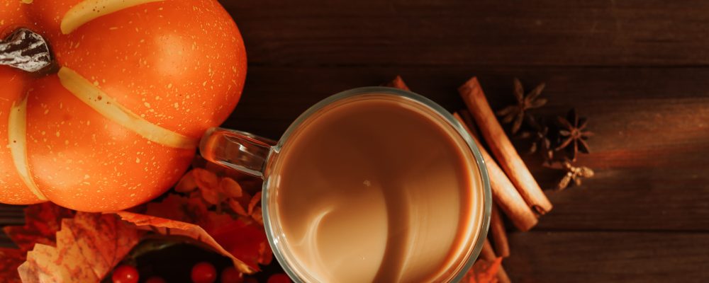 A warm drink sits in a clear glass cup on a wooden table. Nearby are pumpkins and autumn leaves. The scene shows preparation for fall gathering with a cozy feel.