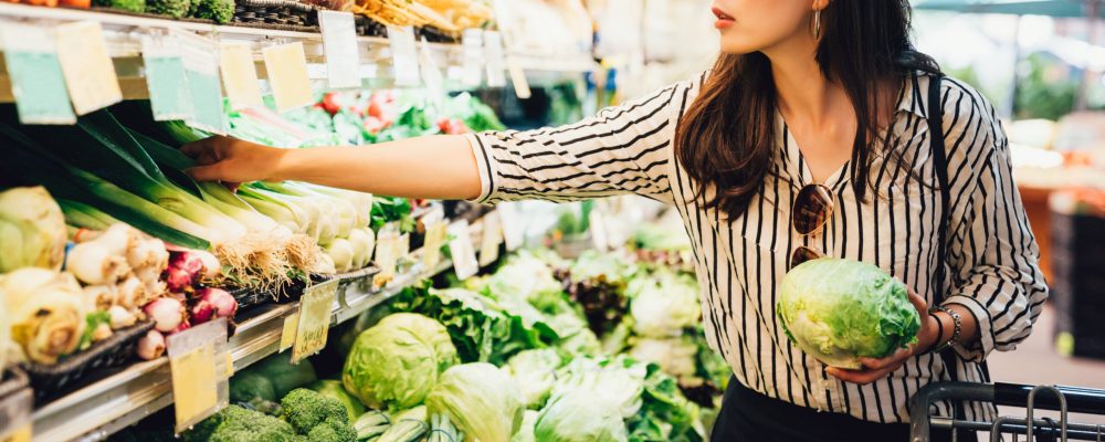 asian local woman buy vegetables and fruits in supermarket. young chinese lady holding green leaf vegetable and picking choosing green onion on cold open refrigerator. elegant female grocery shopping