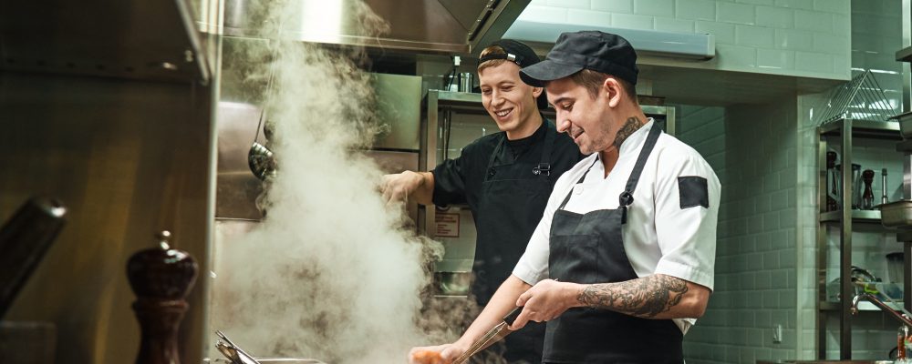 Positive mood. Side view of two cheerful and smiling cooks in uniform are preparing a food in a kitchen restaurant. Food preparation concept