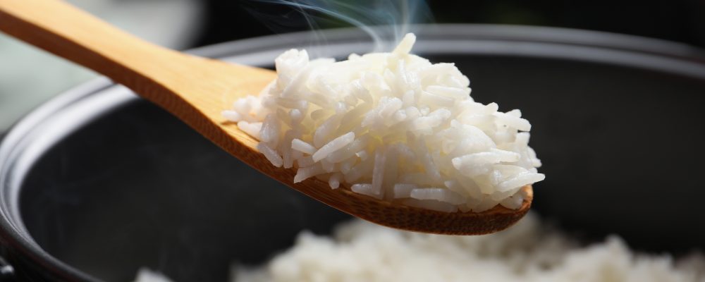 Spoon with tasty hot rice over cooker, closeup
