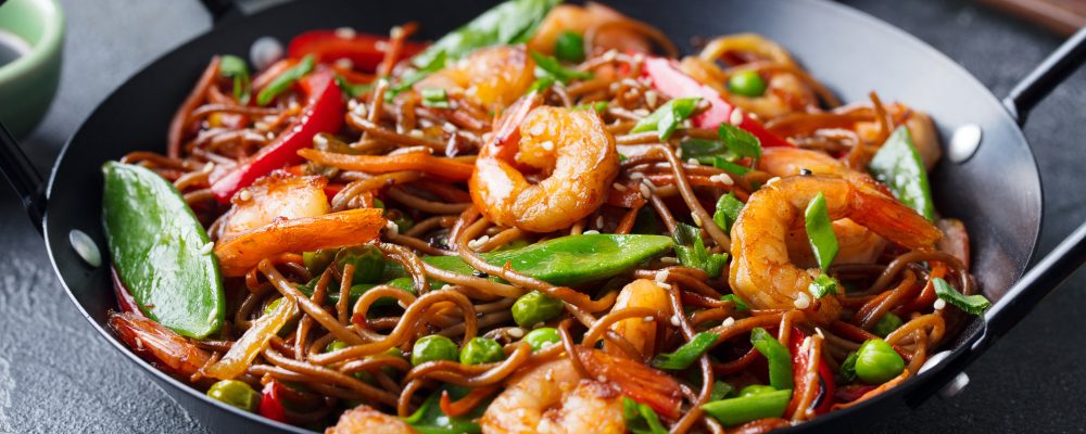 Stir fry noodles with vegetables and shrimps in black iron pan. Slate background. Close up