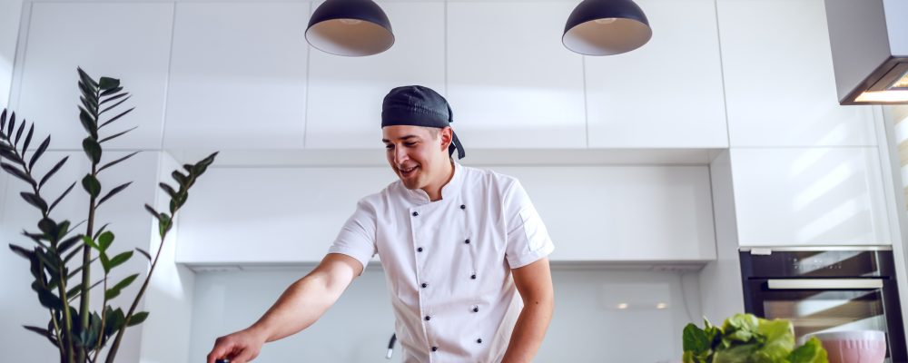 Happy smiling caucasian chef in uniform standing in domestic kitchen and preparing salmon.