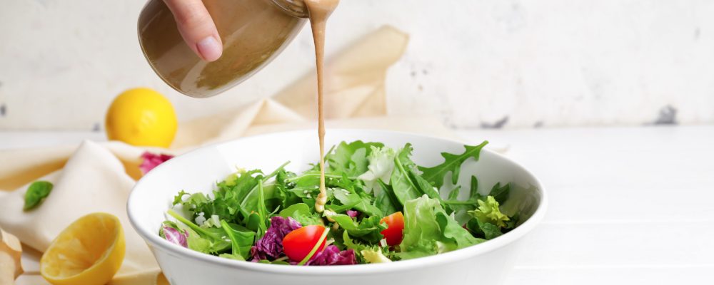 Woman pouring tasty tahini from jar onto vegetable salad in bowl