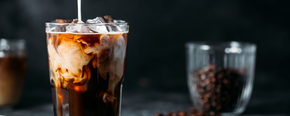 Ice coffee in a tall glass with cream poured over and coffee beans on dark gray background
