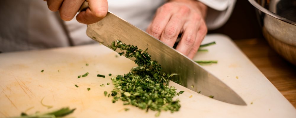 Close-up of hands of chef gently chopping green onion with sharp knife on cutting board