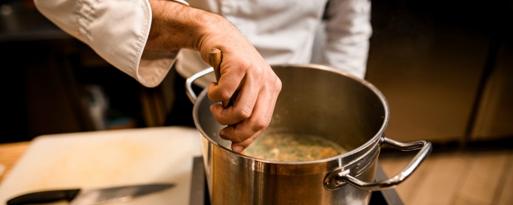 close-up on the hand of male chef in white jacket who is stirring soup in pot with spoon