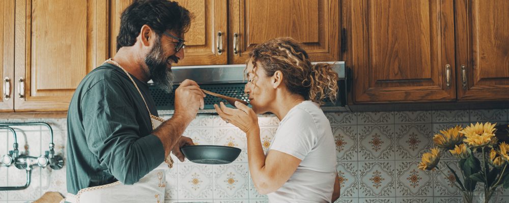 One happy real couple at home enjoying life together and relationship. Man giving food to woman to taste. Male people cooking for his wife. Male and female enjoying leisure activity indoor in kitchen
