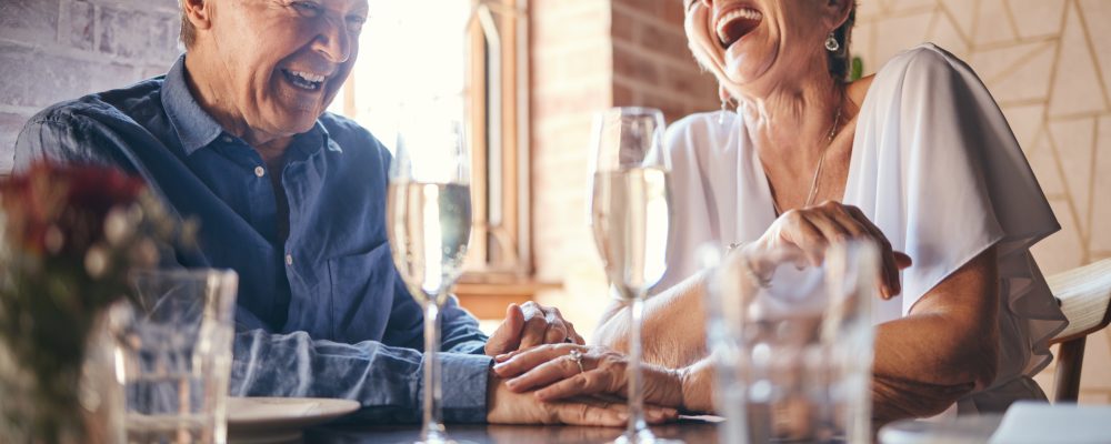Love, laughing and old couple holding hands at a restaurant on a romantic wine date in celebration of a happy marriage. Smile, relaxed and senior woman enjoying glass of champagne with funny partner.