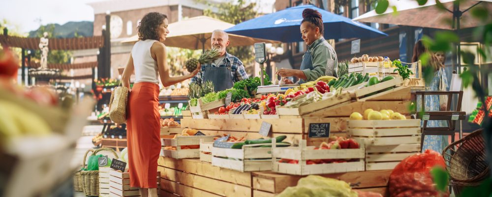 Hispanic Female Customer Buying Two Garlic Heads and a Pineapple From a Multiethnic Farmers Couple. Successful Adults Managing Small Business Farm Stall at a City Square Market