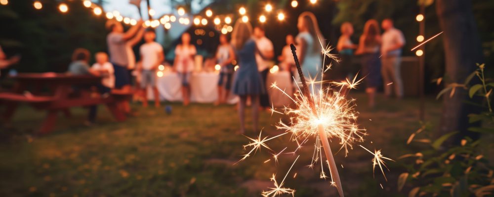 A brightly lit sparkler in the foreground, casting a warm glow, with a festive backyard party in the background. 4th july, memorial. independence