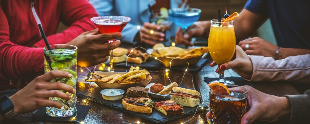 Group of people drinking alcohol and eating appetizers sitting at cocktail bar - Friends enjoying happy hour at brewery pub restaurant - Food and beverage life style concept