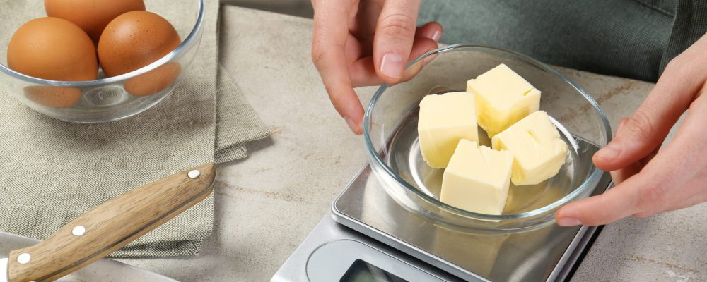 Woman weighing butter on kitchen scale at grey table, closeup